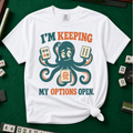 I'm Keeping My Options Open T-Shirt in White, knotted flat lay on a green mahjong mat - worried teal octopus holding a dot tile, a bam tile, and a green dragon tile - "I'M KEEPING MY OPTIONS OPEN." in teal and orange retro letters