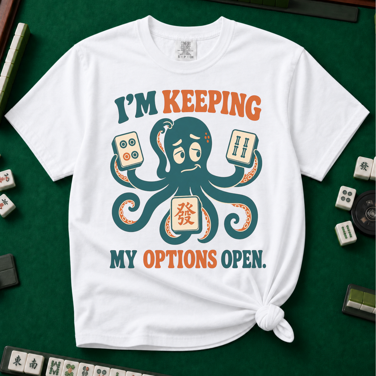 I'm Keeping My Options Open T-Shirt in White, knotted flat lay on a green mahjong mat - worried teal octopus holding a dot tile, a bam tile, and a green dragon tile - "I'M KEEPING MY OPTIONS OPEN." in teal and orange retro letters