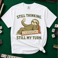 Still Thinking Still My Turn T-Shirt in White, knotted flat lay on a green mahjong mat - a sloth draped along its mahjong tile rack, chin in hand, holding up a red dragon tile - "STILL THINKING. STILL MY TURN." in olive green vintage letters