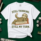Still Thinking Still My Turn T-Shirt in White, knotted flat lay on a green mahjong mat - a sloth draped along its mahjong tile rack, chin in hand, holding up a red dragon tile - "STILL THINKING. STILL MY TURN." in olive green vintage letters
