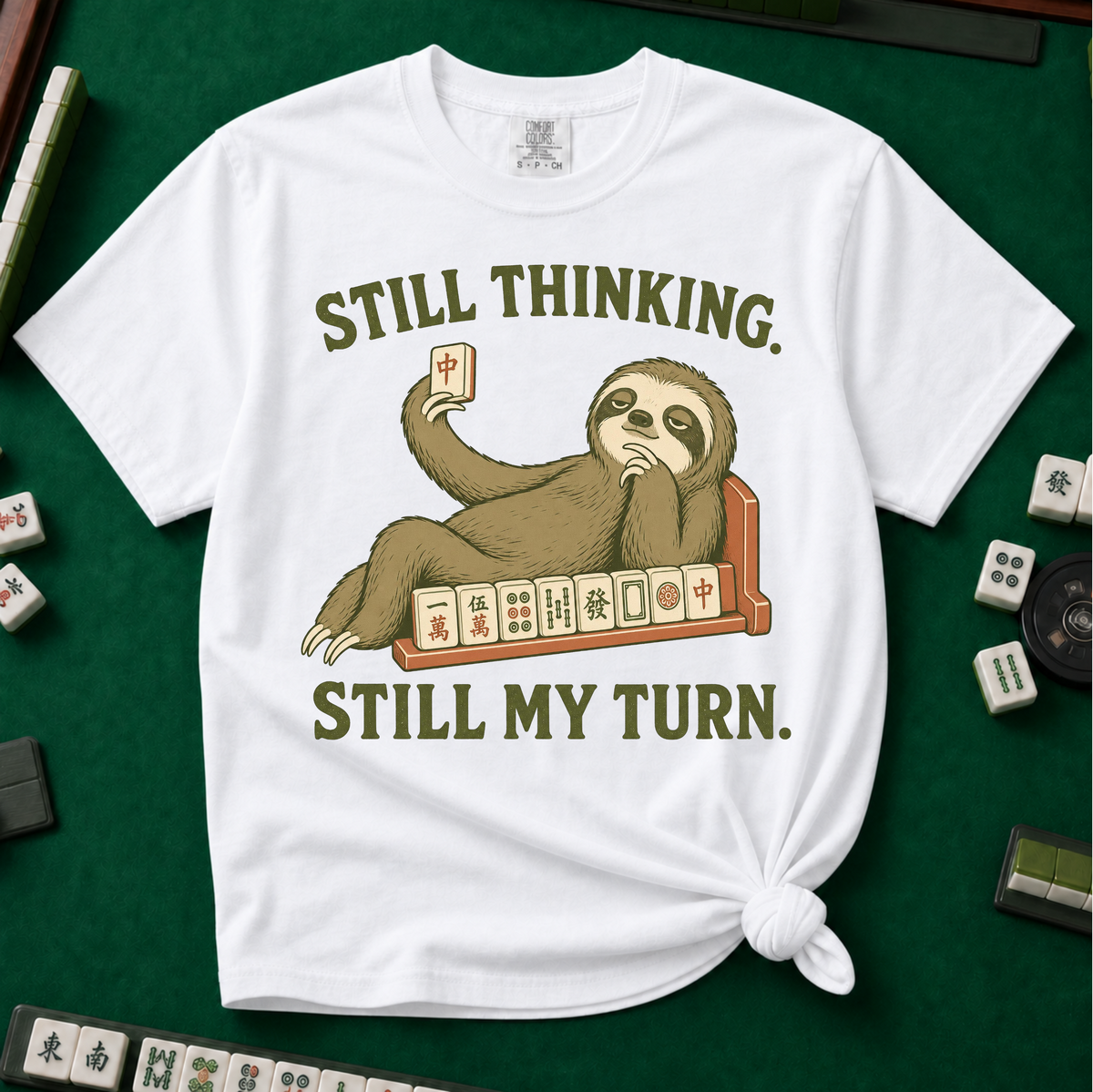 Still Thinking Still My Turn T-Shirt in White, knotted flat lay on a green mahjong mat - a sloth draped along its mahjong tile rack, chin in hand, holding up a red dragon tile - "STILL THINKING. STILL MY TURN." in olive green vintage letters
