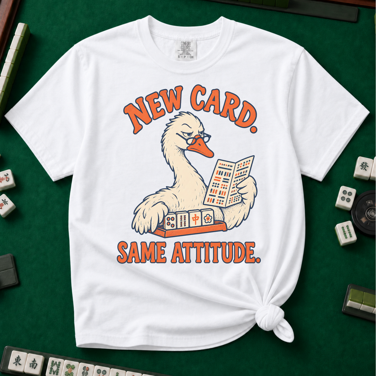 New Card T-Shirt in White - knotted tee flat lay on a green mahjong mat with tiles, printed with a white goose in glasses reading the new mahjong card over a rack of tiles, with the words NEW CARD. SAME ATTITUDE.