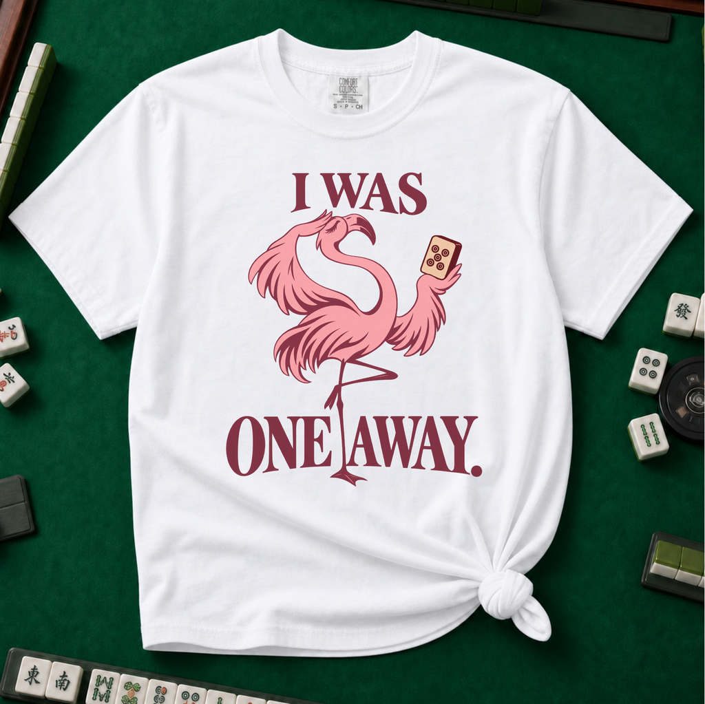 I Was One Away T-Shirt in White - knotted tee flat lay on a green mahjong mat with tiles, printed with a pink flamingo standing on one leg and holding a mahjong tile, with the words I WAS ONE AWAY.