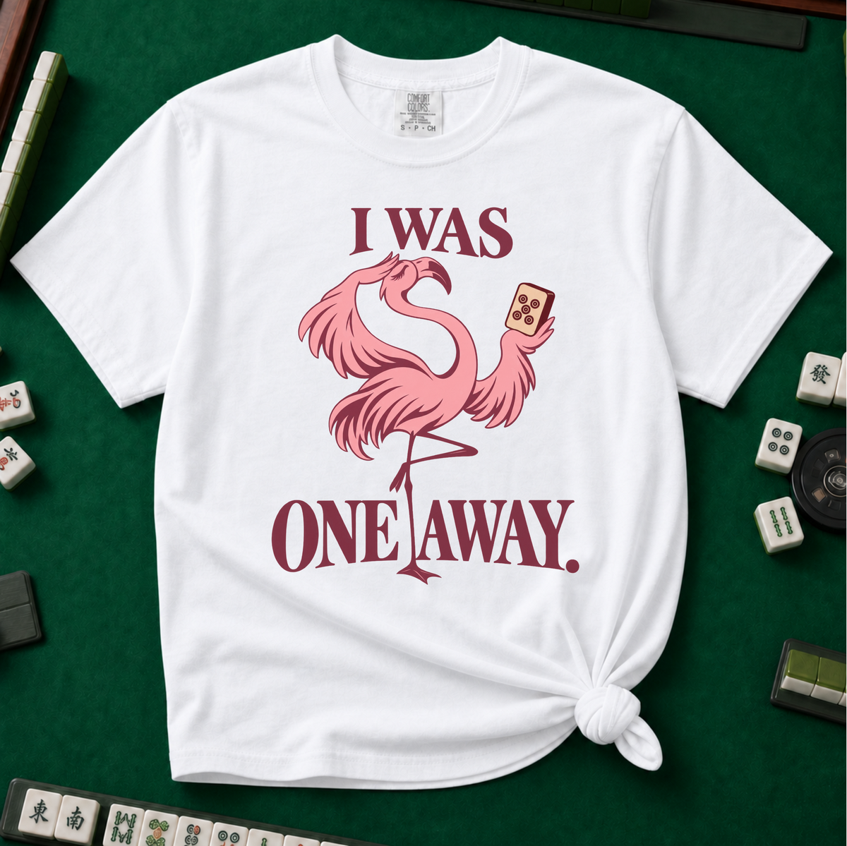 I Was One Away T-Shirt in White - knotted tee flat lay on a green mahjong mat with tiles, printed with a pink flamingo standing on one leg and holding a mahjong tile, with the words I WAS ONE AWAY.