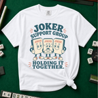 Joker Support Group Holding It Together T-Shirt in White, knotted flat lay on a green mahjong mat - three retro cartoon joker tiles in sneakers, the middle one sweating nervously while the other two hold him up - "JOKER SUPPORT GROUP - HOLDING IT TOGETHER." in teal retro letters