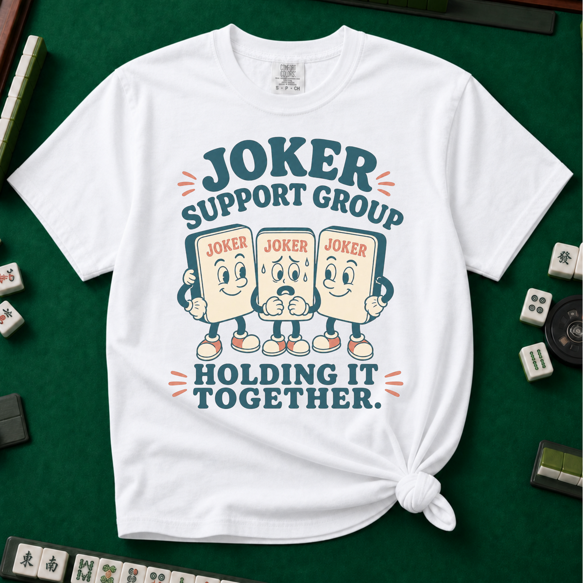Joker Support Group Holding It Together T-Shirt in White, knotted flat lay on a green mahjong mat - three retro cartoon joker tiles in sneakers, the middle one sweating nervously while the other two hold him up - "JOKER SUPPORT GROUP - HOLDING IT TOGETHER." in teal retro letters