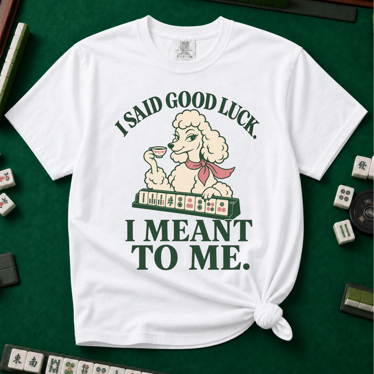 I Said Good Luck T-Shirt in White - knotted tee flat lay on a green mahjong mat with tiles, printed with a cream poodle in a pink scarf holding a teacup behind a rack of mahjong tiles, with the words I SAID GOOD LUCK. I MEANT TO ME.