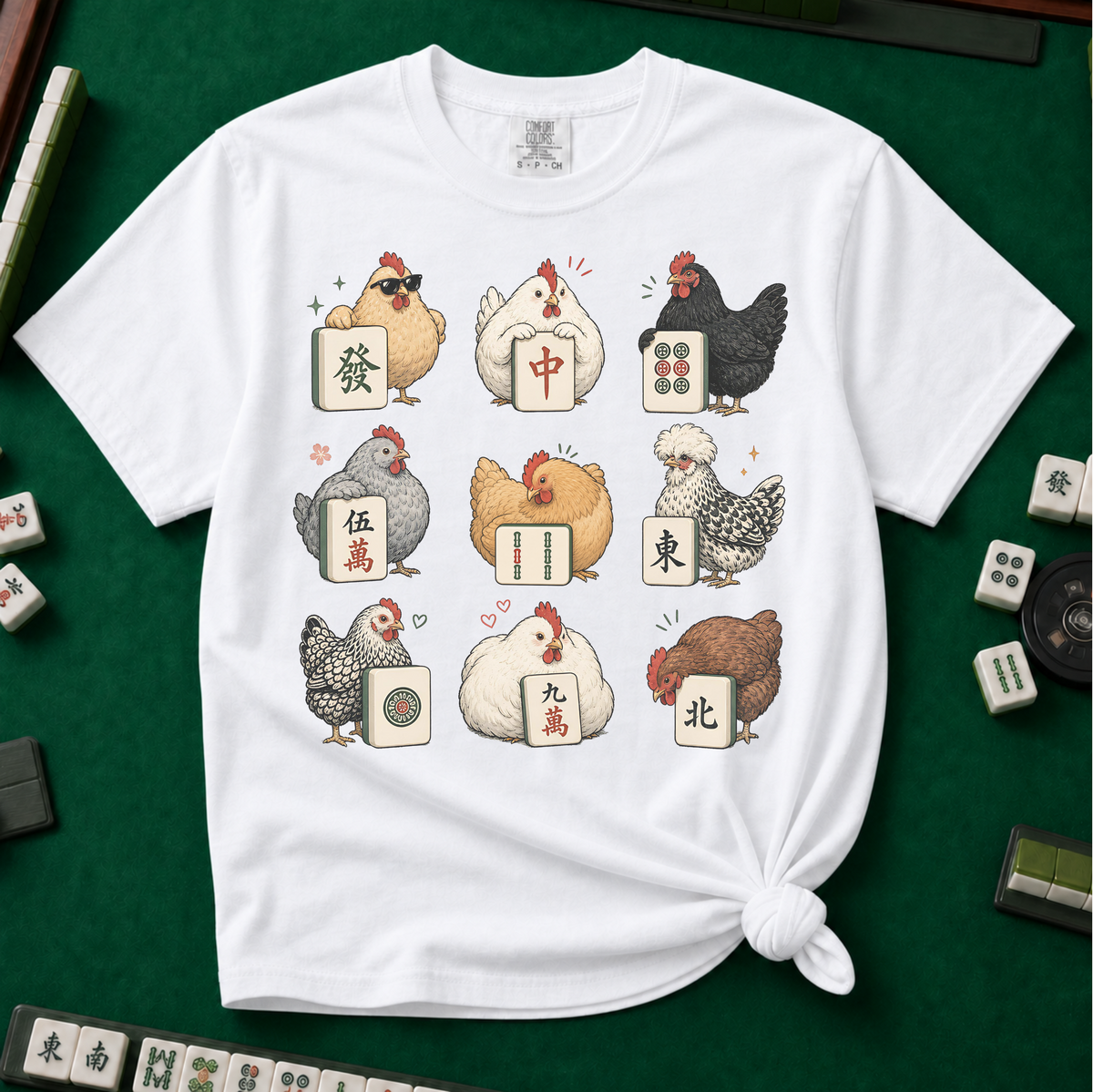 Mahjong Chickens T-Shirt in White, knotted flat lay on a green mahjong mat - a 3x3 grid of nine hens, each posing with a mahjong tile - a sunglasses hen with the green dragon, a white hen with the red dragon, a black hen with dots, a crested hen with East, and more