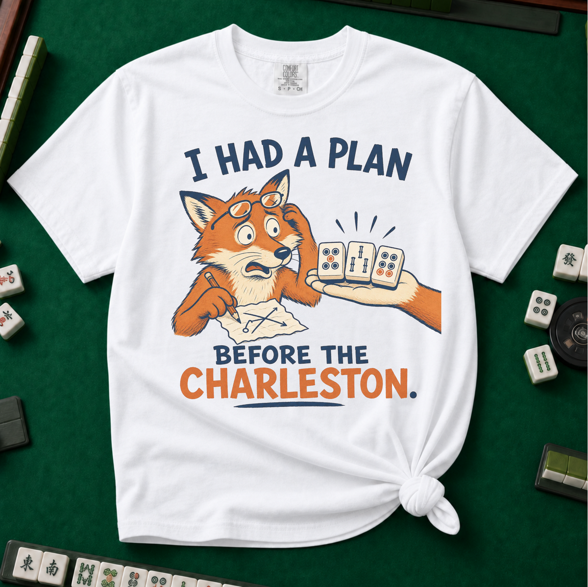 I Had A Plan Before The Charleston T-Shirt in White, knotted flat lay on a green mahjong mat - a stressed cartoon fox with glasses and a strategy sketch being handed three mahjong tiles, with the words "I HAD A PLAN BEFORE THE CHARLESTON."