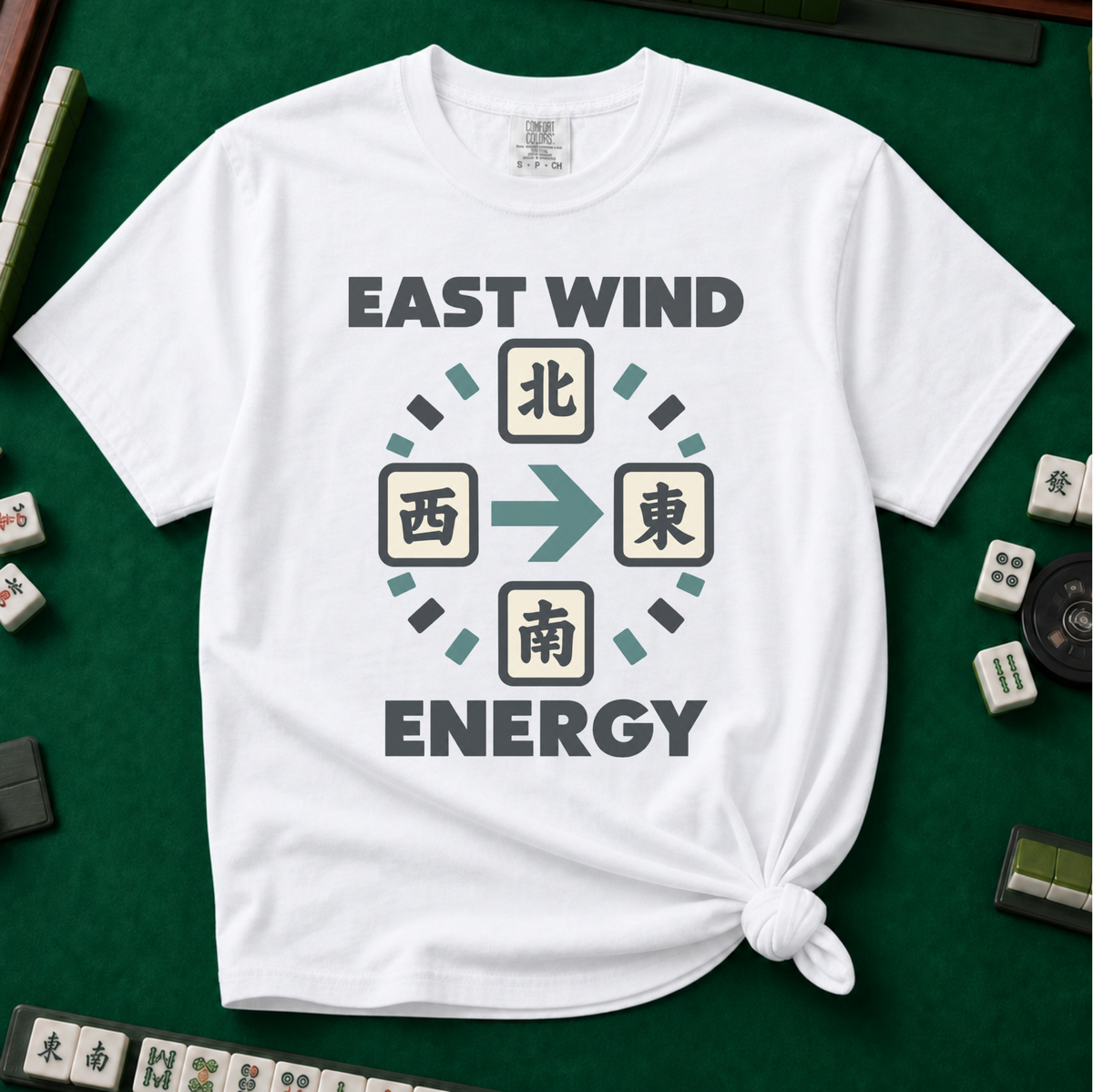 East Wind Energy T-Shirt in White, knotted flat lay on a green mahjong mat - the four mahjong wind tiles arranged in a compass circle - North on top, West left, East right, South below - with a big teal arrow pointing East, between "EAST WIND" and "ENERGY" in charcoal block letters