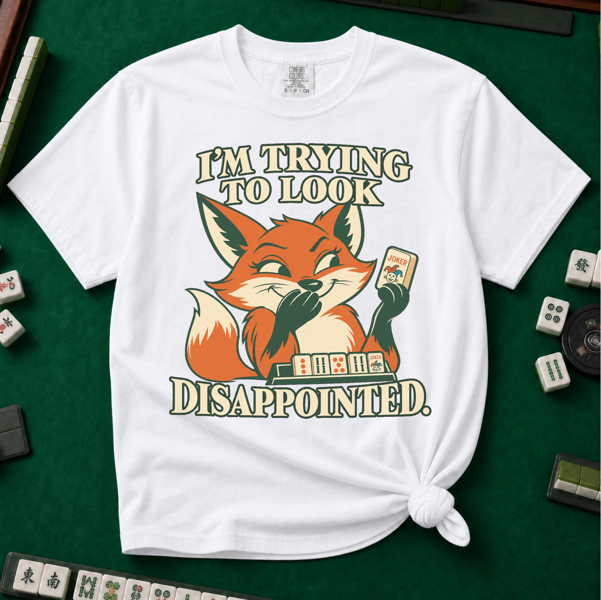 I'm Trying To Look Disappointed T-Shirt in White, knotted flat lay on a green mahjong mat - smug retro fox holding up a joker tile, hiding a grin behind one paw, tile rack in front - "I'M TRYING TO LOOK DISAPPOINTED." in cream collegiate letters