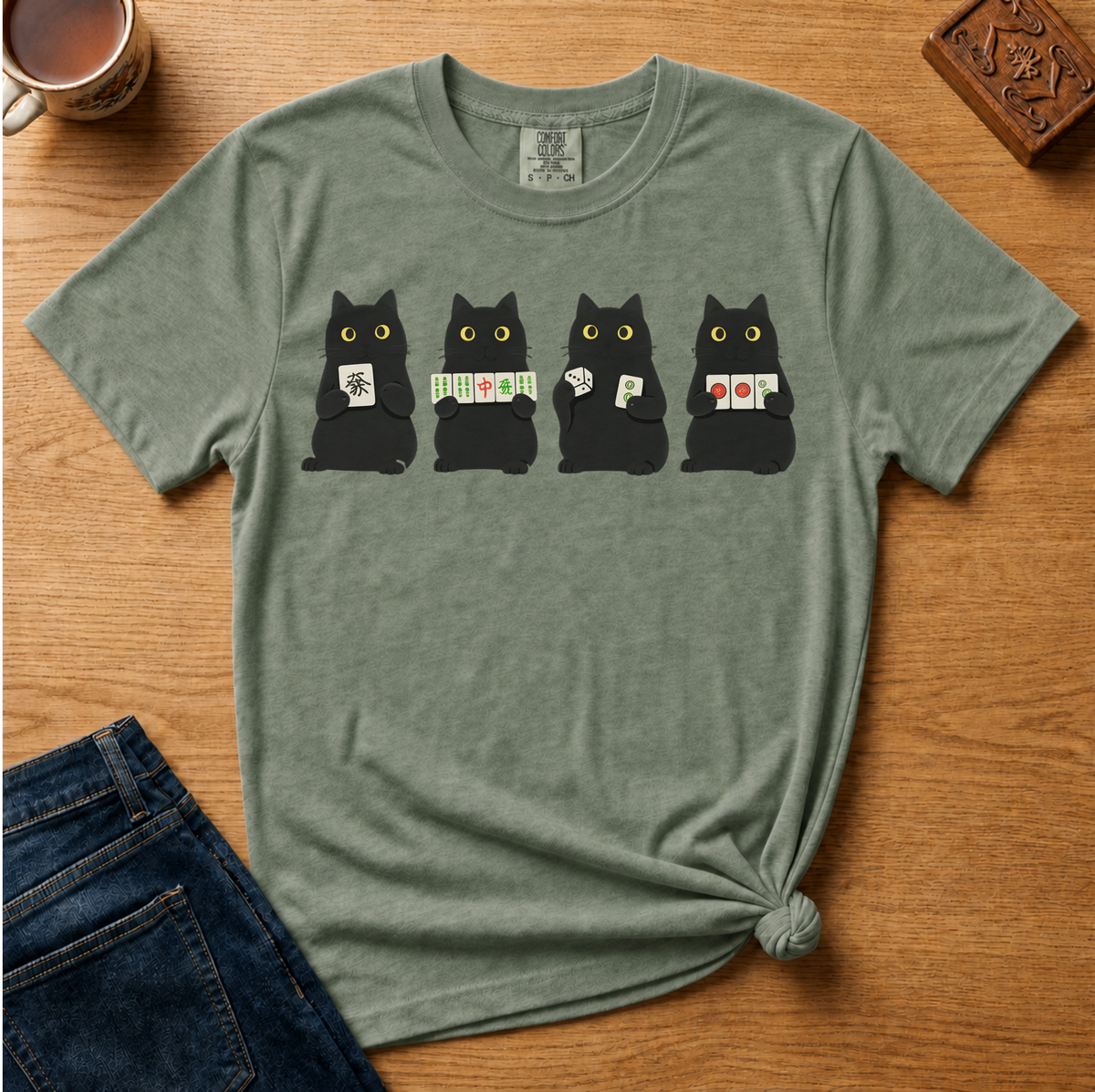 A sage green graphic t-shirt laid flat on a wooden table, styled with a pair of blue jeans, a coffee mug, and a wooden block. The t-shirt features a graphic of four black cats holding playing cards, with the middle two cats wearing green and white striped scarves.