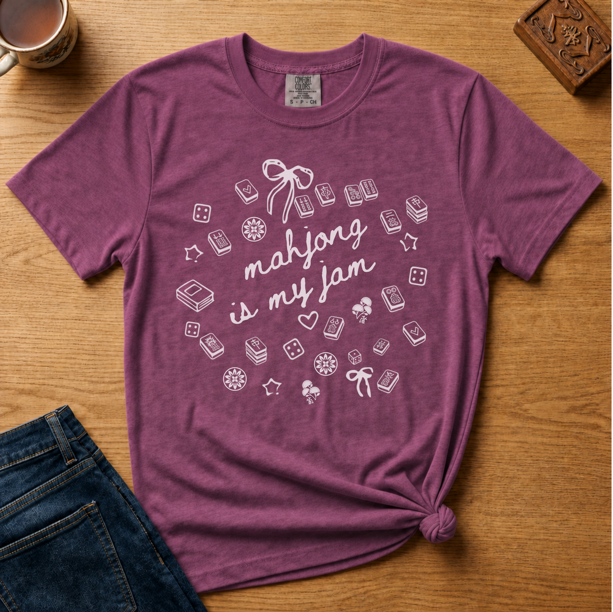 A heathered purple graphic t-shirt with 'mahjong is my jam' printed in white script, surrounded by small white line-art illustrations of mahjong tiles and a bow. The t-shirt is styled in a flat-lay on a wooden table next to a pair of blue jeans, a ceramic mug, and a carved wooden block.