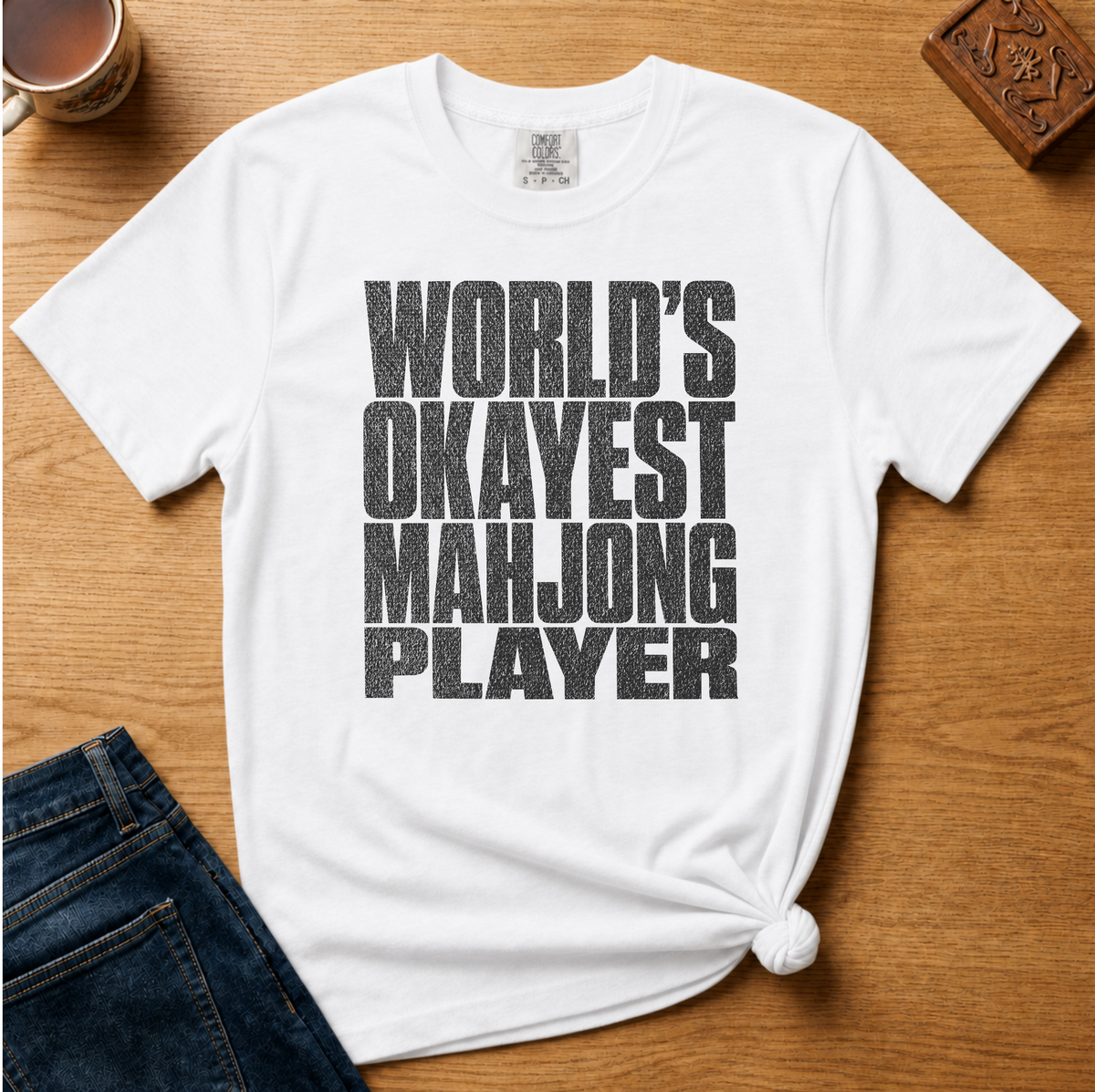 A white short-sleeve graphic T-shirt laid flat on a wooden surface, styled with dark blue jeans at the bottom left and a mug and carved wooden block as props. The shirt has large distressed black text reading “WORLD’S OKAYEST MAHJONG PLAYER” across the front.