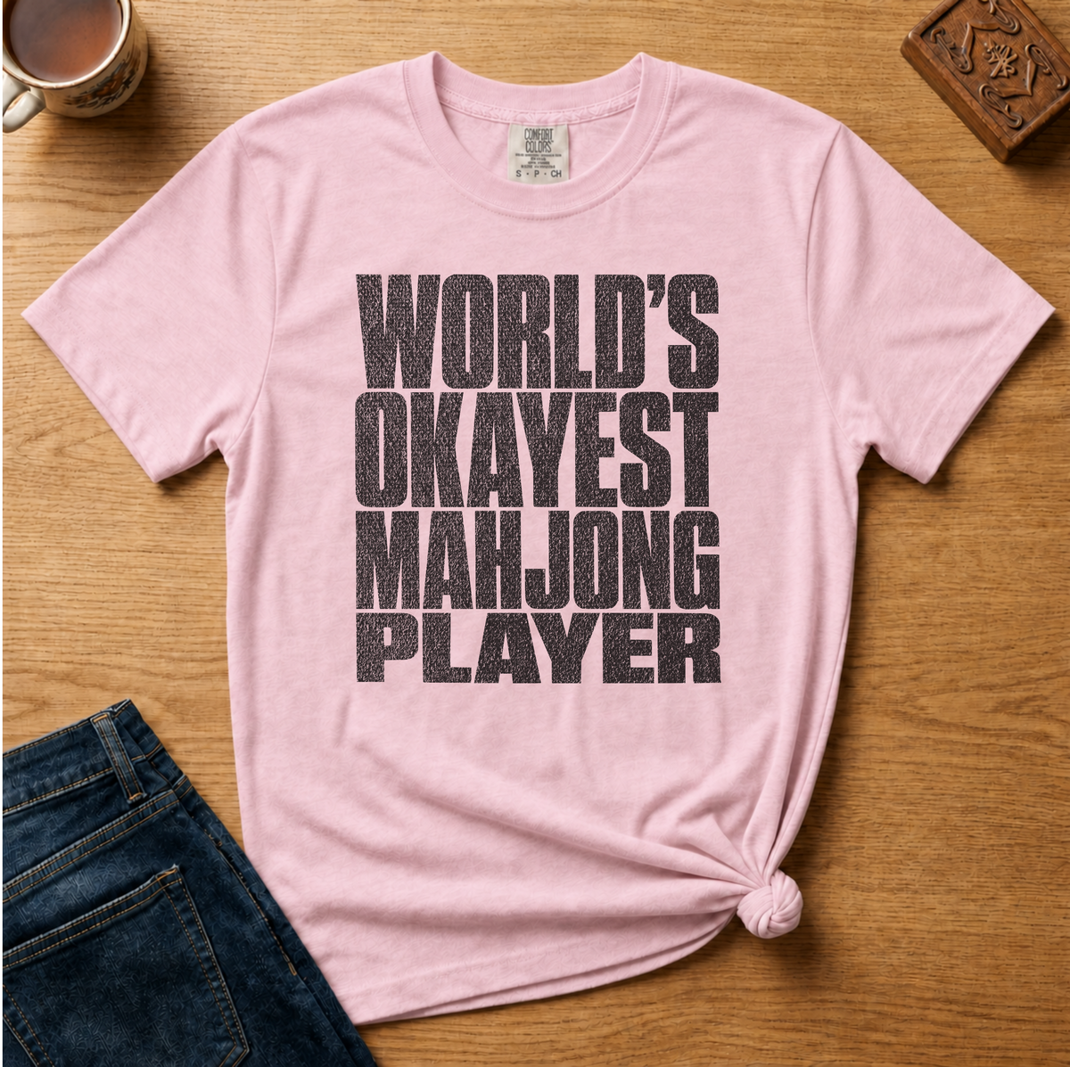 A light pink short-sleeve t-shirt with a distressed black text graphic reading 'WORLD'S OKAYEST MAHJONG PLAYER' laid flat on a wooden surface, styled with dark blue jeans, a coffee cup, and a carved wooden block.