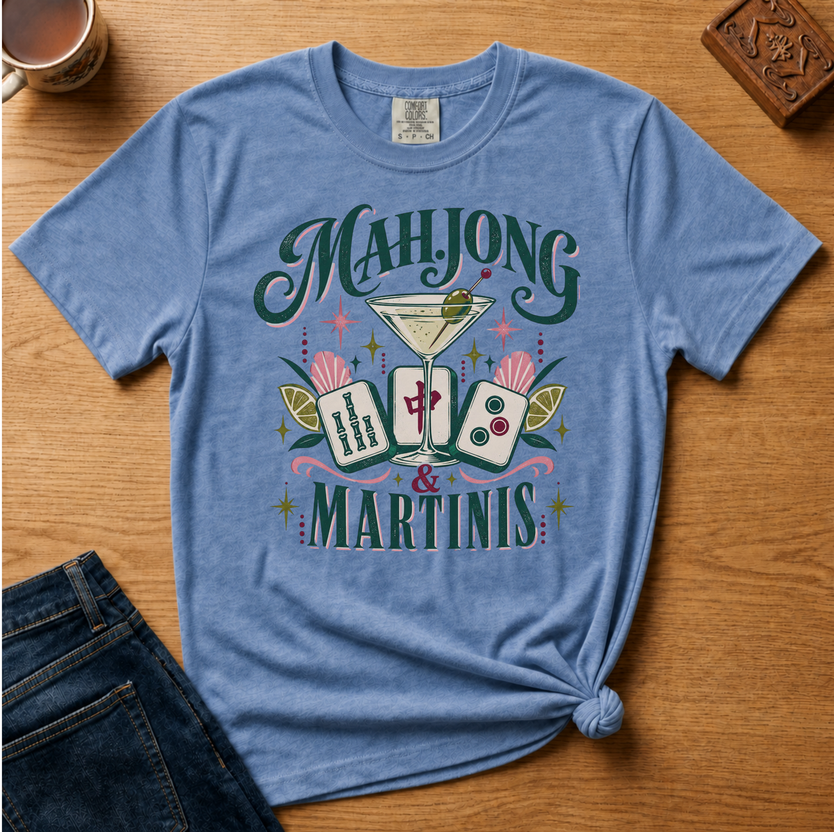 A light blue graphic t-shirt laid flat on a wooden table, styled with a denim jacket, a mug, and a small carved wooden block. The t-shirt features a colorful graphic with the words 'MAHJONG & MARTINIS' and illustrations of playing cards, a martini glass, and pink palm leaves.