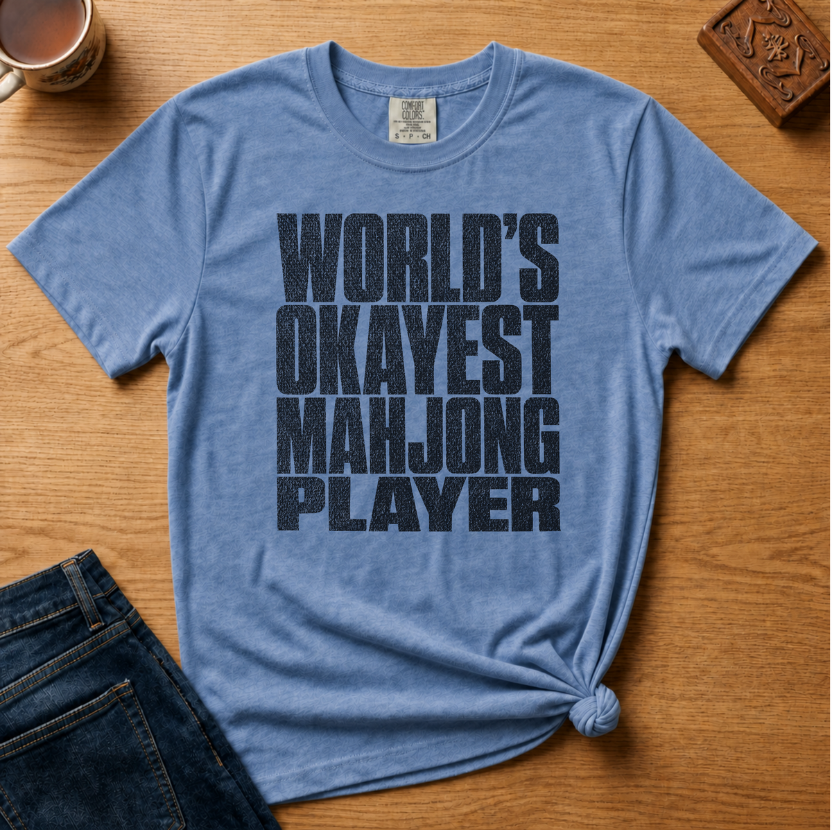A heathered blue short-sleeve t-shirt with the text 'WORLD'S OKAYEST MAHJONG PLAYER' printed in distressed black lettering. The shirt is styled in a flat lay on a wooden table with a denim jeans leg, a coffee mug, and a carved wooden block as props.