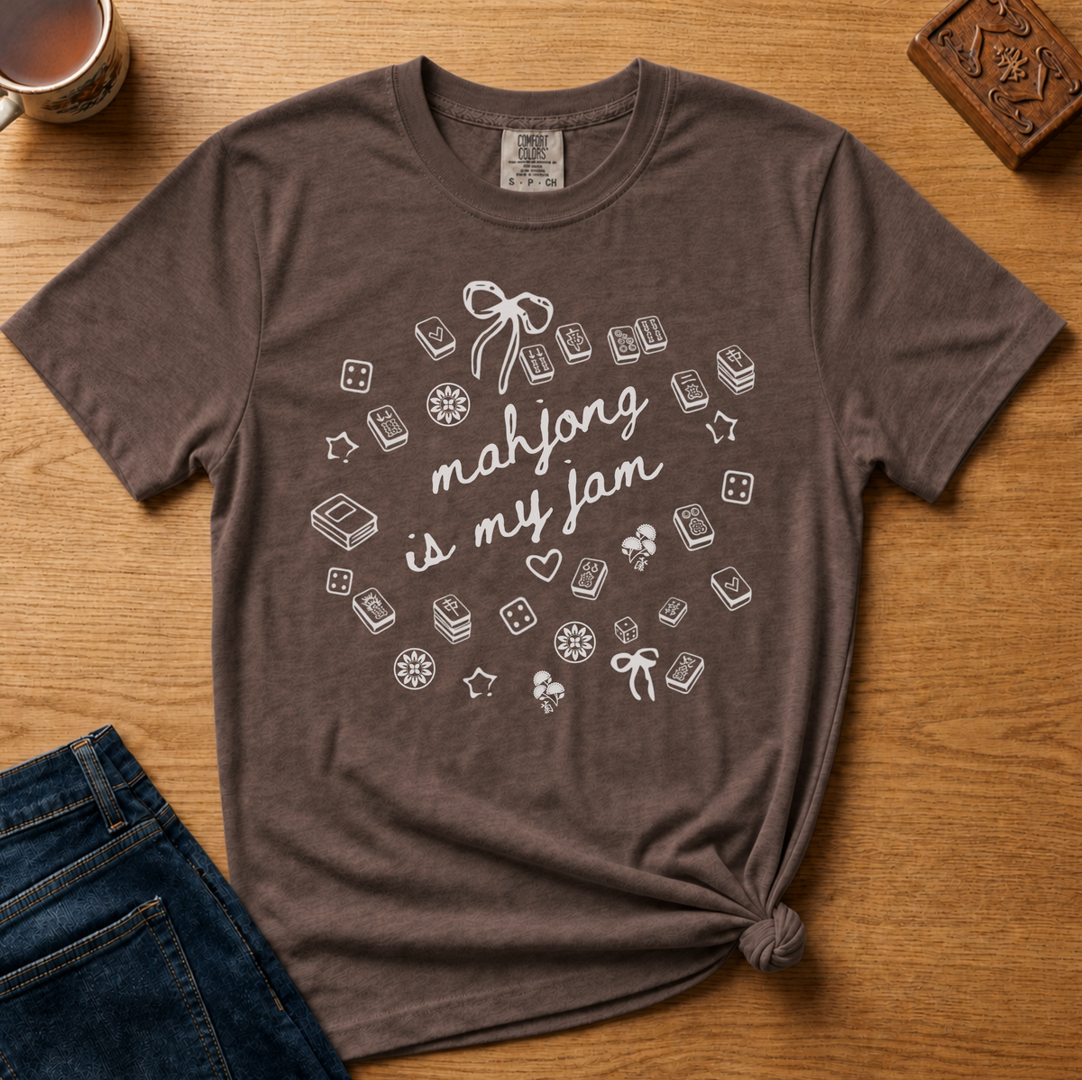 A brown short-sleeve graphic T-shirt laid flat on a wooden surface, styled with a mug, phone, and denim shorts nearby. The shirt has a white printed design featuring the phrase 'mahjong is my jam' surrounded by small game-related icons.