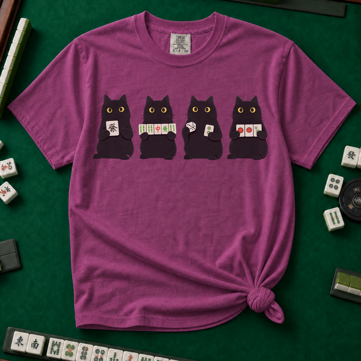 Black Cat Mahjong Shirt in Berry - knotted tee on a green mahjong mat with scattered tiles, printed with Mahjong Social Club lettering and four black cats peeking over the words holding tiles