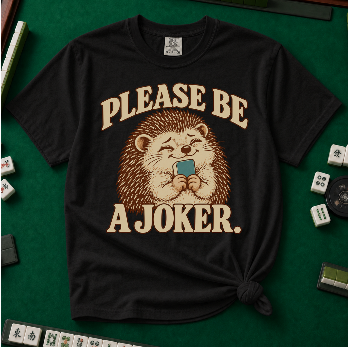 Please Be A Joker Shirt in Black - tee laid flat on a green mahjong mat with scattered tiles, printed with a hedgehog wearing a pink bow and a little crown and the words Please Be A Joker