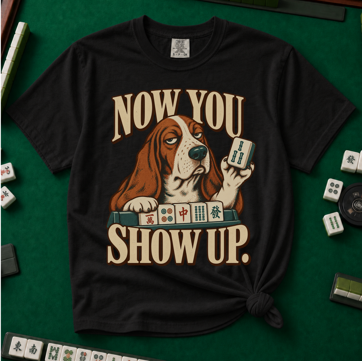 Now You Show Up Shirt in Black - knotted tee on a green mahjong mat with scattered tiles, printed with a basset hound peeking over a row of mahjong tiles and the words Now You Show Up