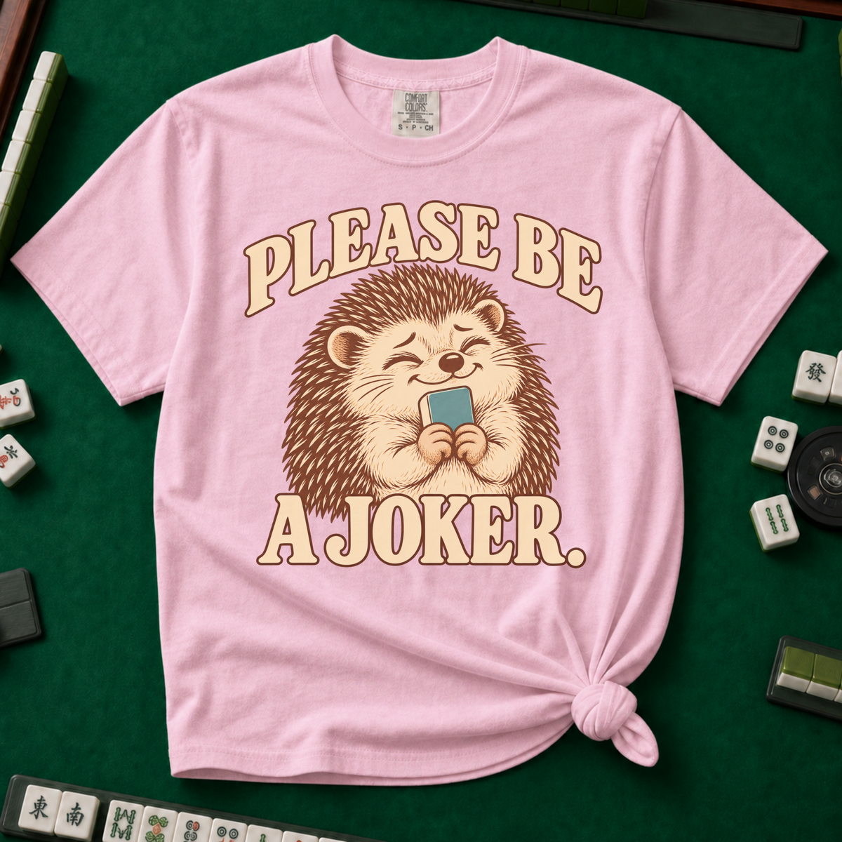 Please Be A Joker Shirt in Blossom - knotted tee on a green mahjong mat with scattered tiles, printed with a hedgehog wearing a pink bow and a little crown and the words Please Be A Joker