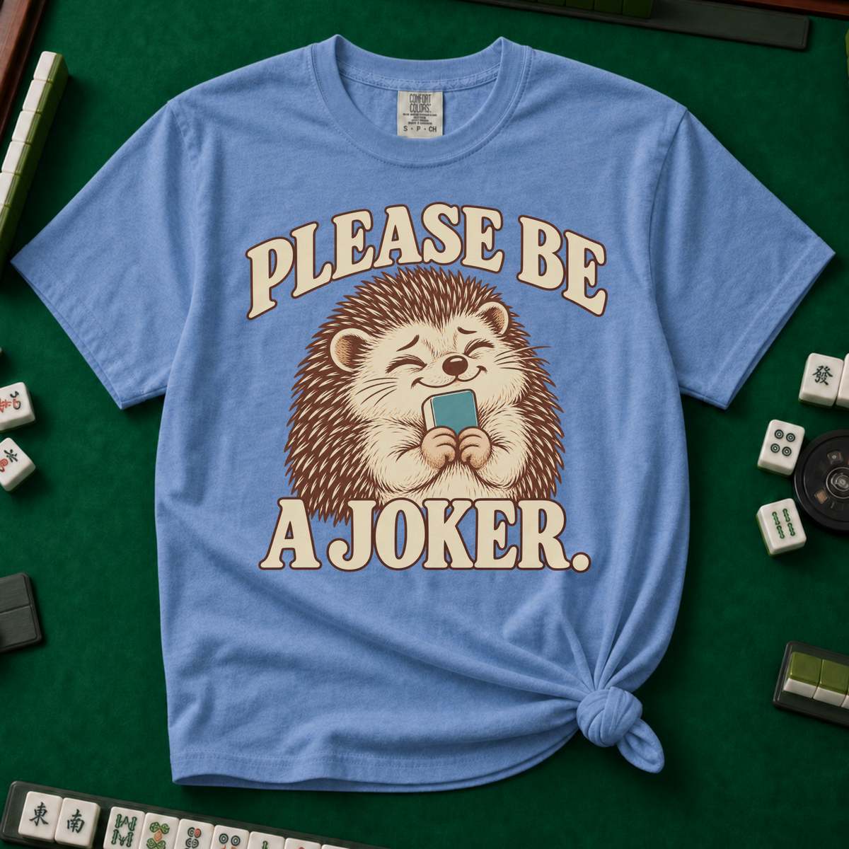 Please Be A Joker Shirt in Blue Jean - knotted tee on a green mahjong mat with scattered tiles, printed with a hedgehog wearing a pink bow and a little crown and the words Please Be A Joker
