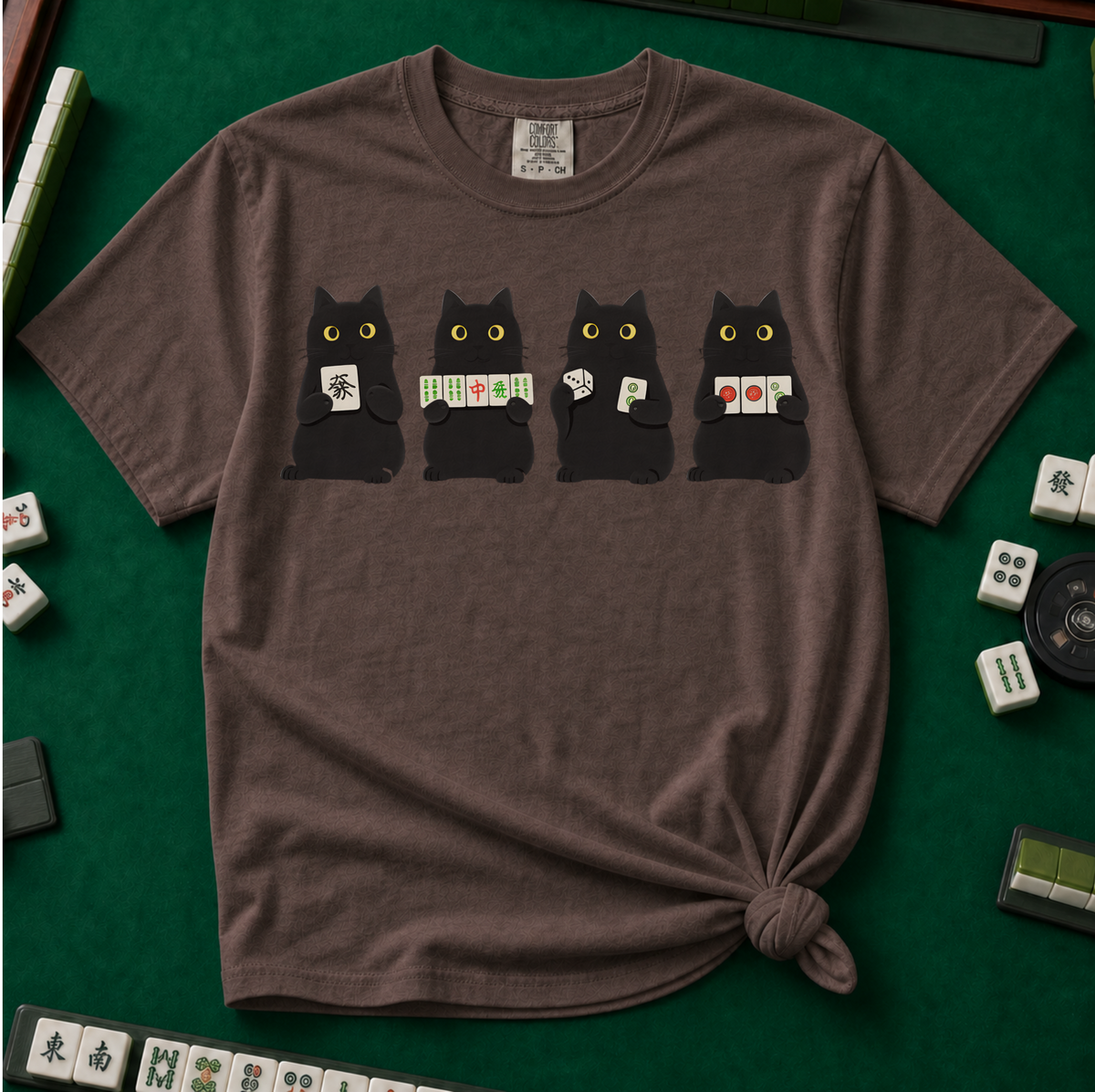 Black Cat Mahjong Shirt in Espresso - tee laid flat on a green mahjong mat with scattered tiles, printed with American Mahjong Social Club lettering and four black cats holding mahjong tiles