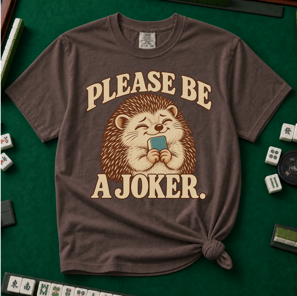Please Be A Joker Shirt in Espresso - knotted tee on a green mahjong mat with scattered tiles, printed with a hedgehog wearing a pink bow and a little crown and the words Please Be A Joker