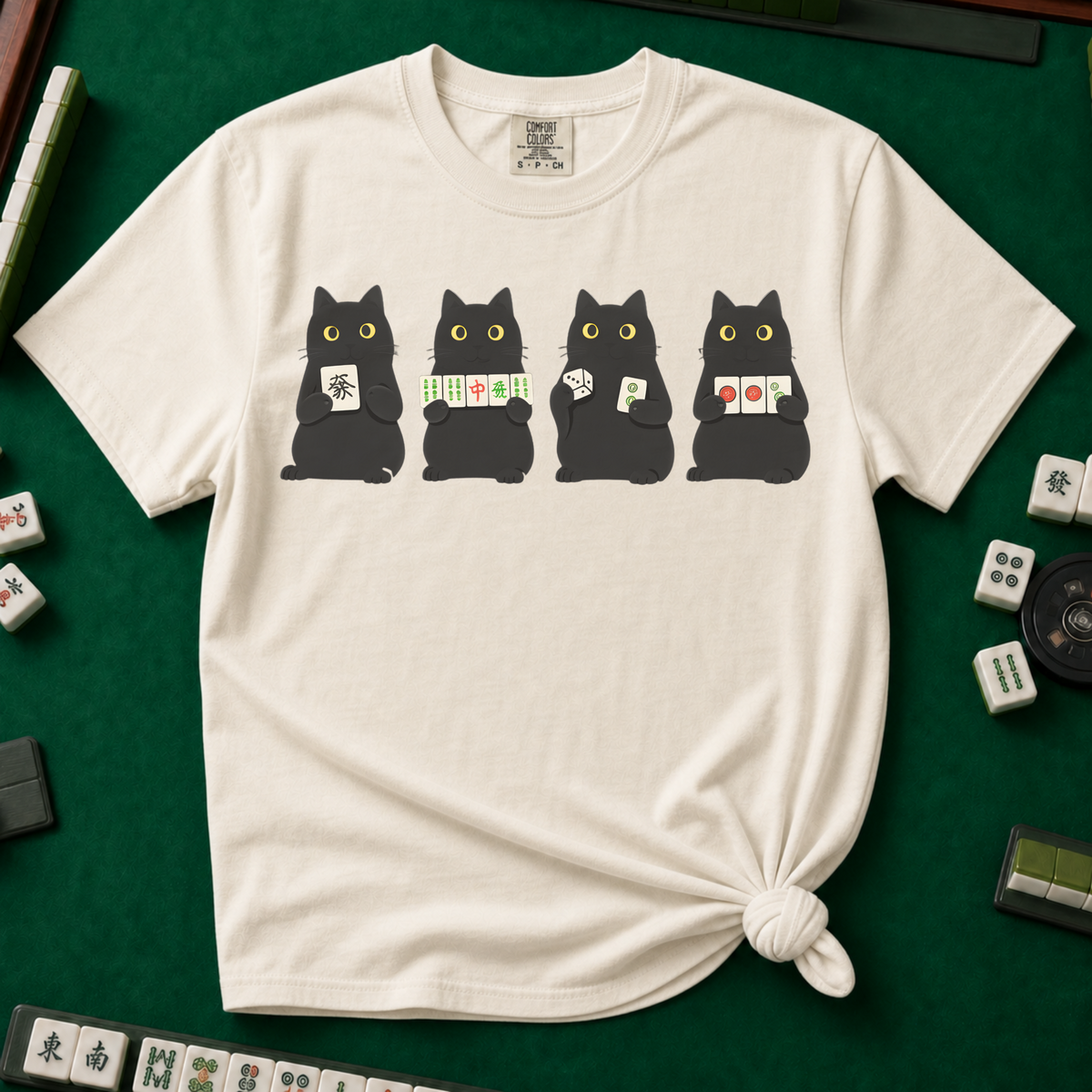 Black Cat Mahjong Shirt in Ivory - knotted tee on a green mahjong mat with scattered tiles, printed with Mahjong Social Club lettering and four black cats peeking over the words holding tiles