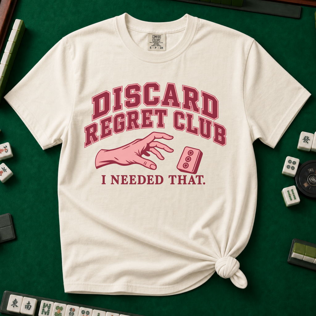 Discard Regret Club I Needed That T-Shirt in Ivory, knotted flat lay on a green mahjong mat - burgundy varsity letters "DISCARD REGRET CLUB" over a pink hand reaching back for a pink dot tile it just discarded - "I NEEDED THAT."