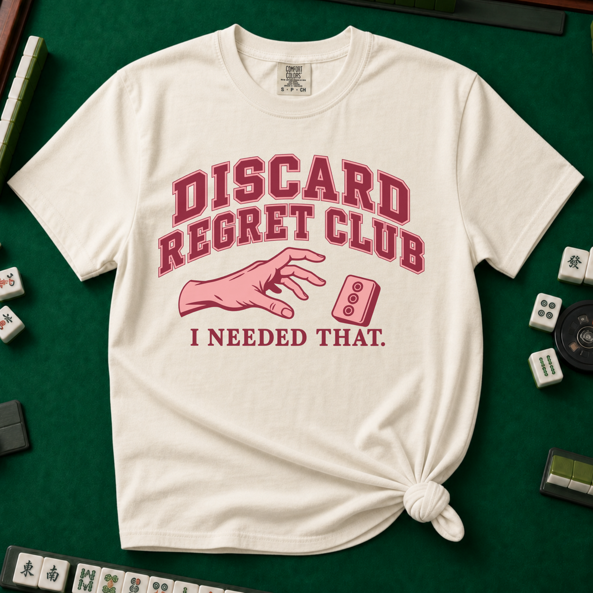 Discard Regret Club I Needed That T-Shirt in Ivory, knotted flat lay on a green mahjong mat - burgundy varsity letters "DISCARD REGRET CLUB" over a pink hand reaching back for a pink dot tile it just discarded - "I NEEDED THAT."