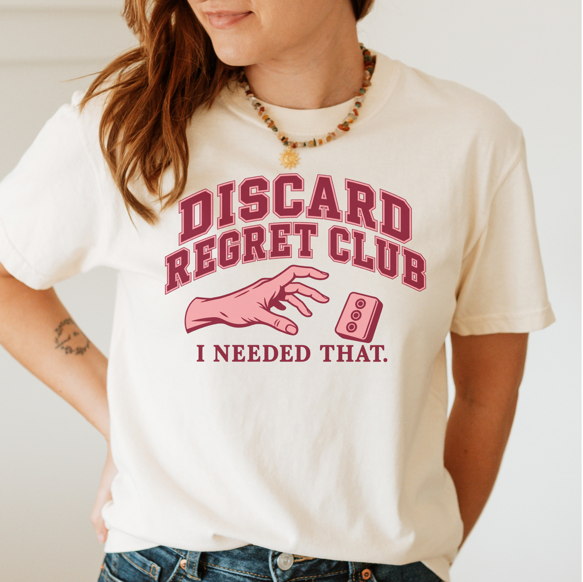 Woman wearing the Discard Regret Club I Needed That T-Shirt in Ivory - burgundy varsity letters "DISCARD REGRET CLUB" over a pink hand reaching back for a pink dot tile it just discarded - "I NEEDED THAT."