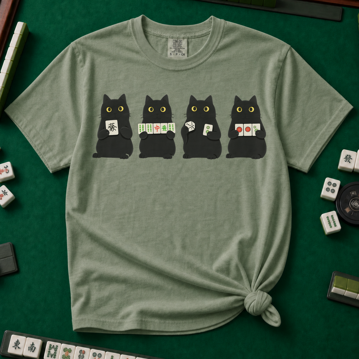 Black Cat Mahjong Shirt in Moss - knotted tee on a green mahjong mat with scattered tiles, printed with American Mahjong Social Club lettering and four black cats holding mahjong tiles