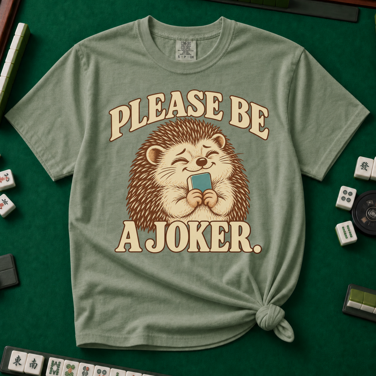 Please Be A Joker Shirt in Moss - knotted tee on a green mahjong mat with scattered tiles, printed with a hedgehog wearing a pink bow and a little crown and the words Please Be A Joker