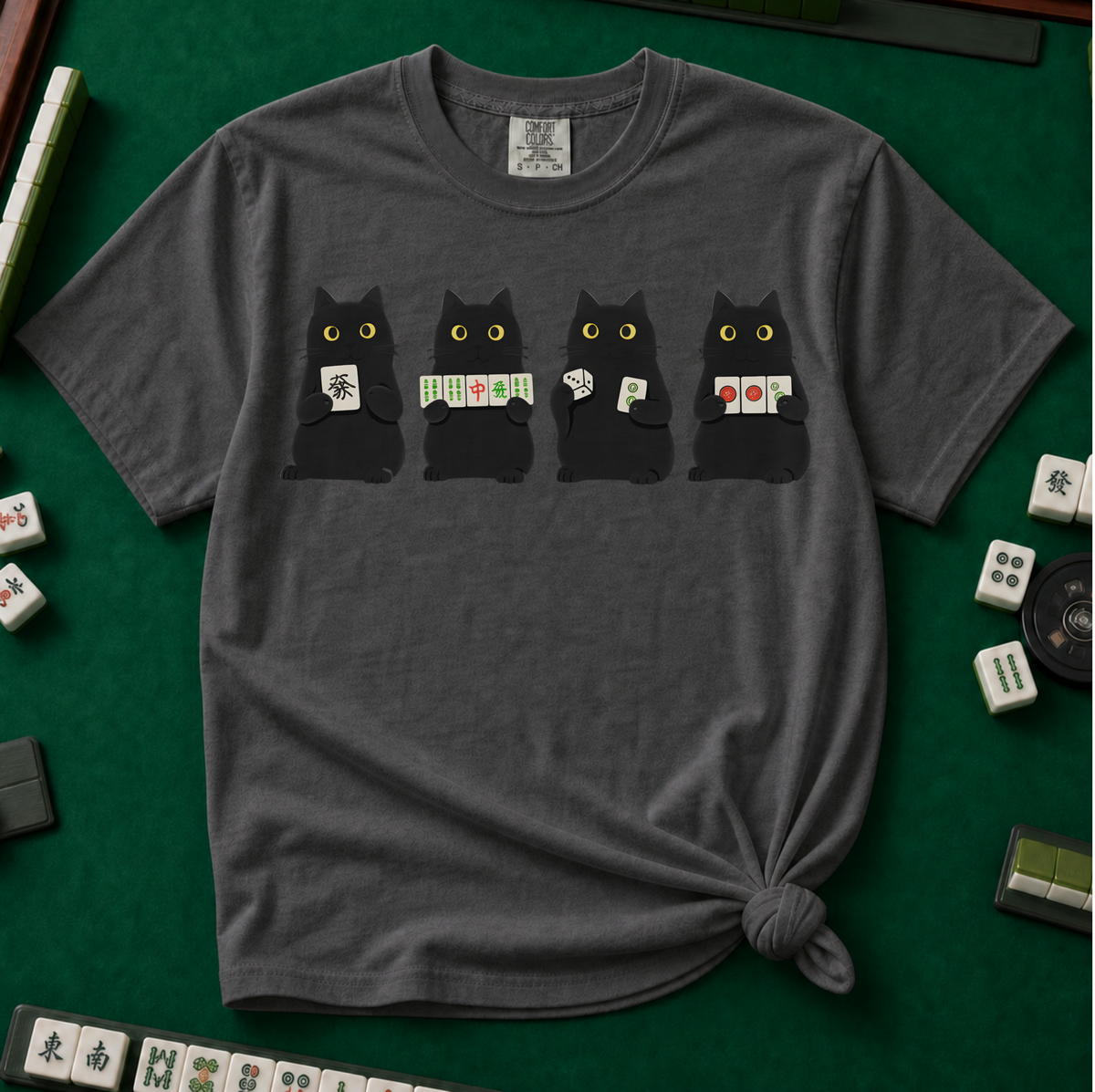 Black Cat Mahjong Shirt in Pepper - knotted tee on a green mahjong mat with scattered tiles, printed with American Mahjong Social Club lettering and four black cats holding mahjong tiles