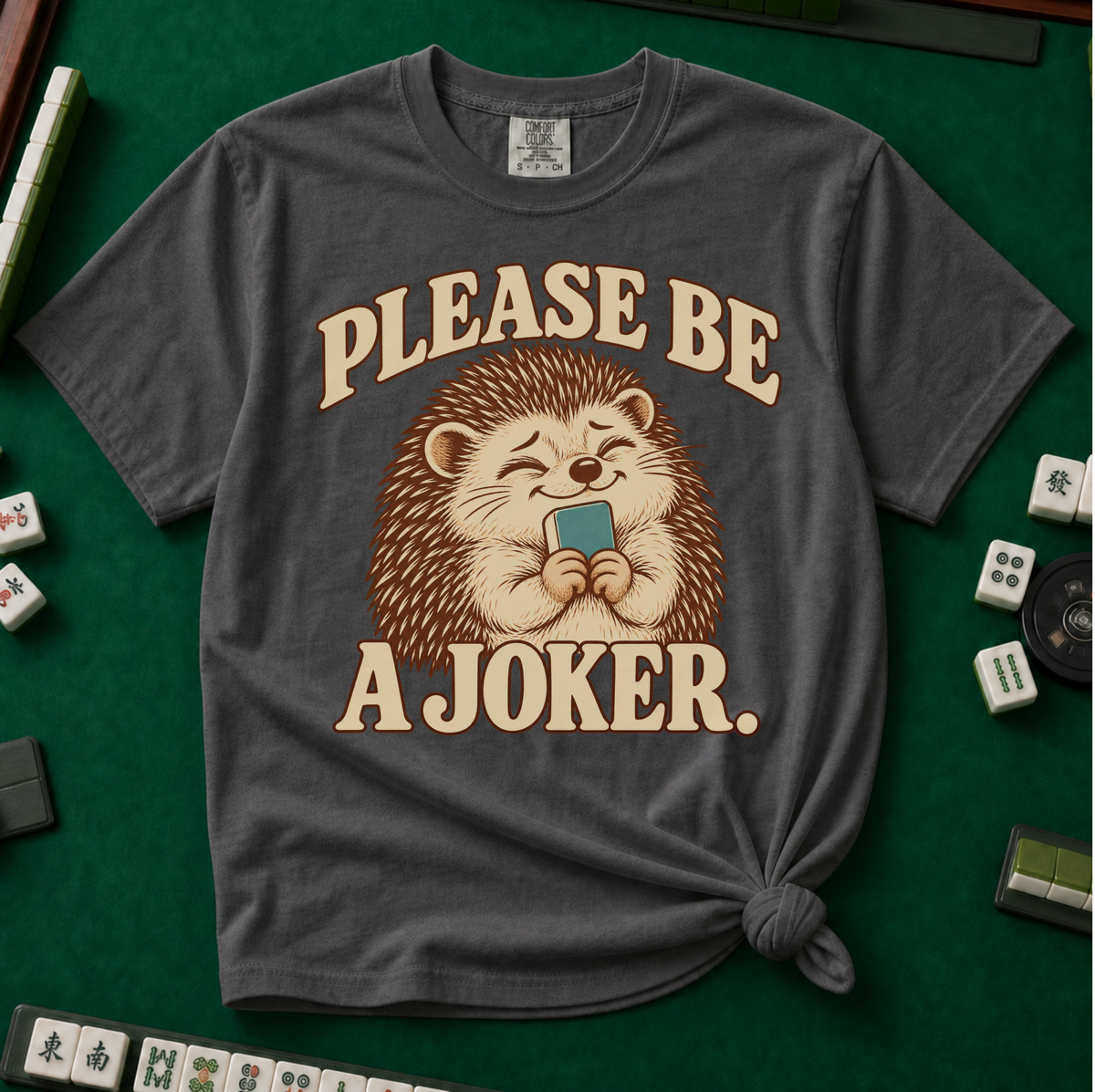 Please Be A Joker Shirt in Pepper - tee laid flat on a green mahjong mat with scattered tiles, printed with a hedgehog wearing a pink bow and a little crown and the words Please Be A Joker