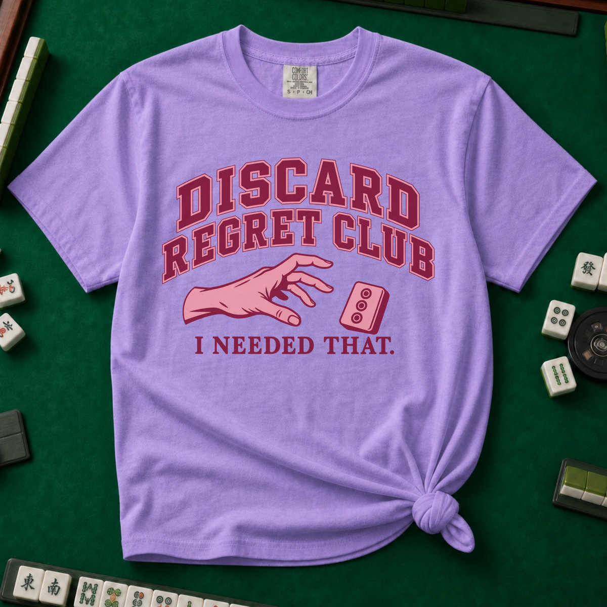 Discard Regret Club I Needed That T-Shirt in Violet, knotted flat lay on a green mahjong mat - burgundy varsity letters "DISCARD REGRET CLUB" over a pink hand reaching back for a pink dot tile it just discarded - "I NEEDED THAT."