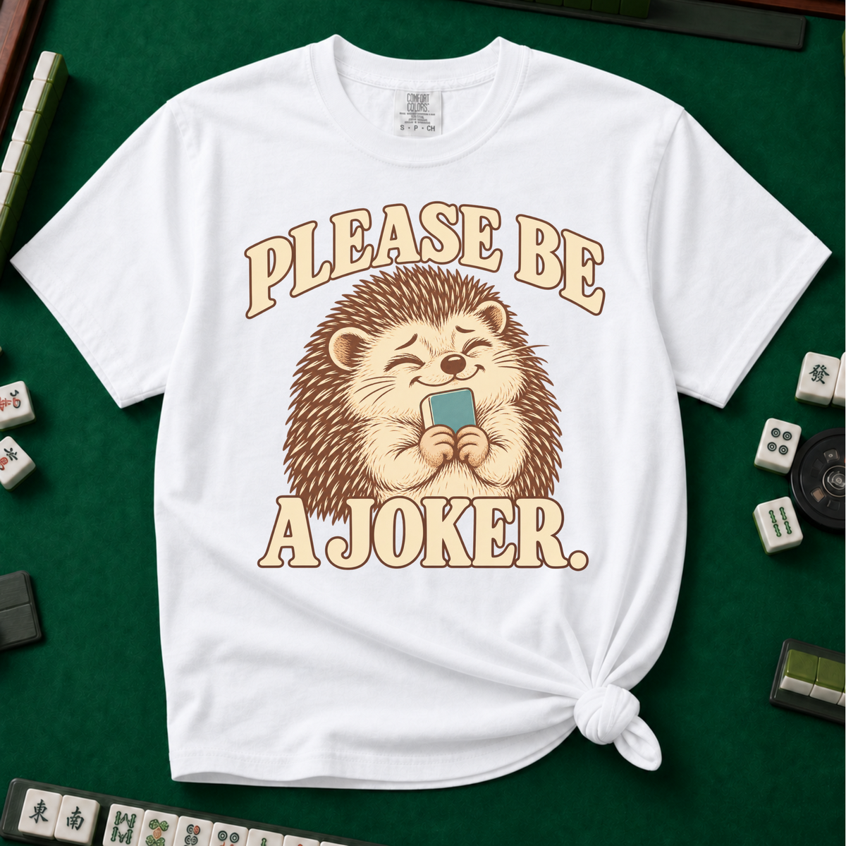 Please Be A Joker Shirt in White - knotted tee on a green mahjong mat with scattered tiles, printed with a hedgehog wearing a pink bow and a little crown and the words Please Be A Joker