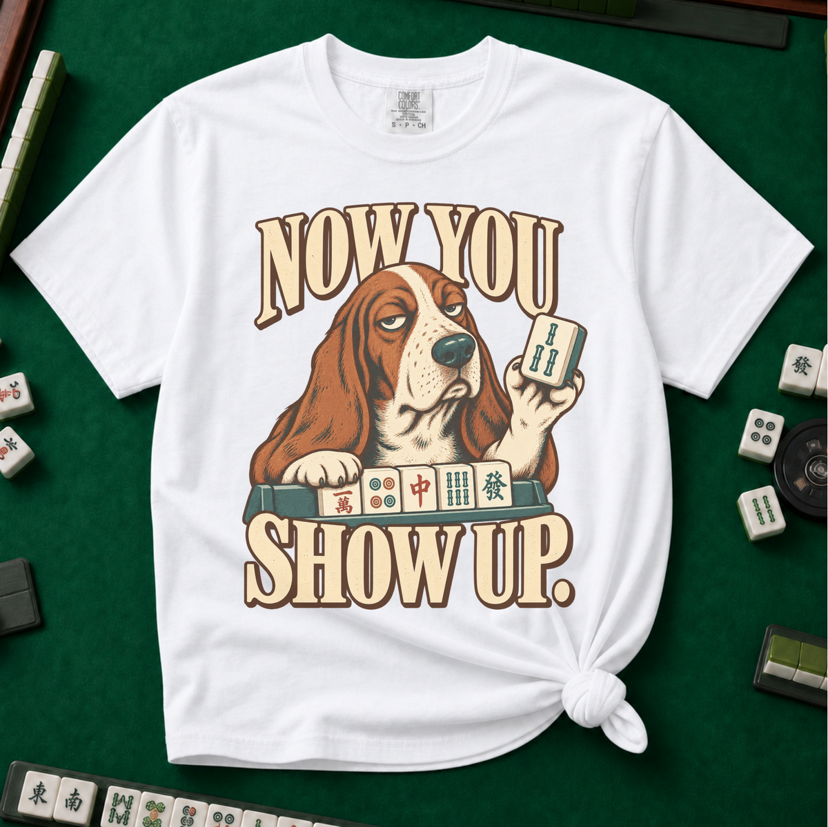 Now You Show Up Shirt in White - knotted tee on a green mahjong mat with scattered tiles, printed with a basset hound peeking over a row of mahjong tiles and the words Now You Show Up