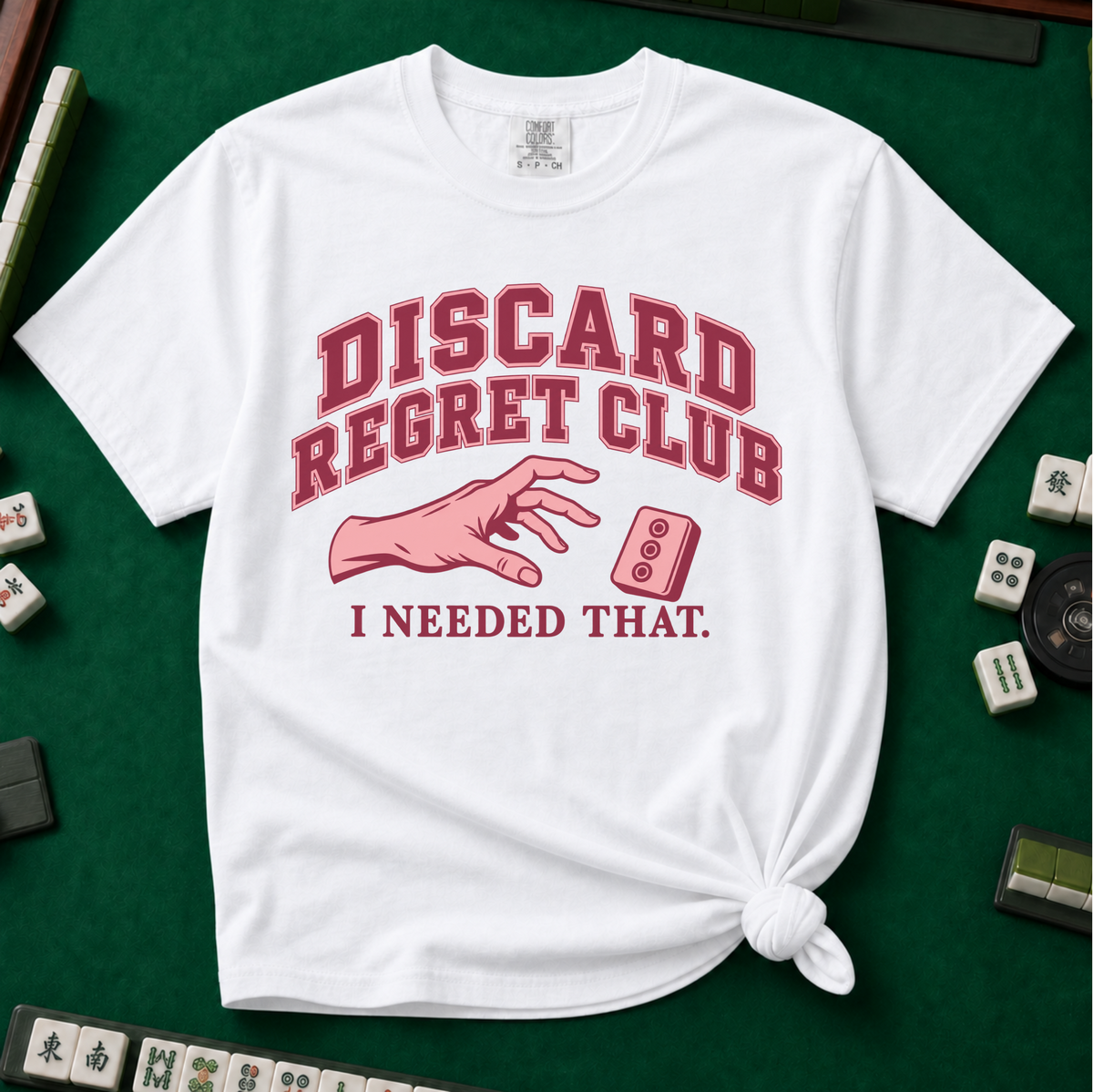 Discard Regret Club I Needed That T-Shirt in White, knotted flat lay on a green mahjong mat - burgundy varsity letters "DISCARD REGRET CLUB" over a pink hand reaching back for a pink dot tile it just discarded - "I NEEDED THAT."