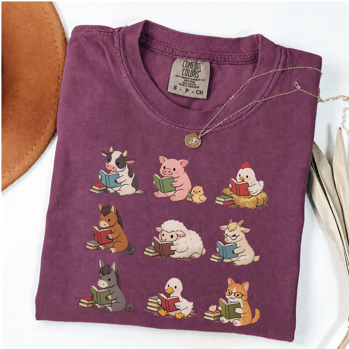 A folded purple t-shirt with a cartoon graphic of farm animals reading books, styled on a white background with a gold necklace and a brown hat edge.