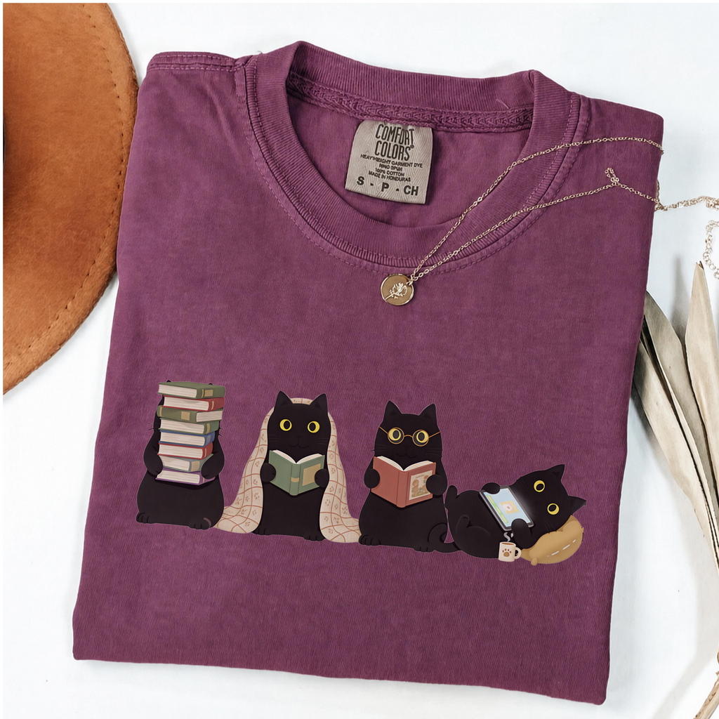 Four black cats with books, a blanket, glasses and an e-reader on a berry shirt.