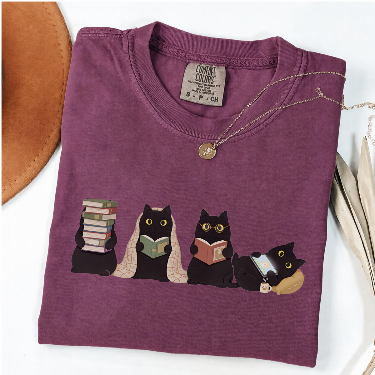 Four black cats with books, a blanket, glasses and an e-reader on a berry shirt.