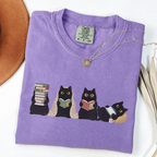 Four black cats with books, a blanket, glasses and an e-reader on a violet shirt.