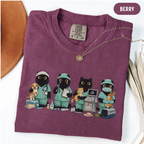 A folded berry purple t-shirt from Comfort Colors, featuring a graphic of four black cats dressed in green veterinary scrubs with their pets. The shirt is styled on a white surface with a gold necklace draped over the collar, a brown leather object on the left, and a beige straw hat on the right.