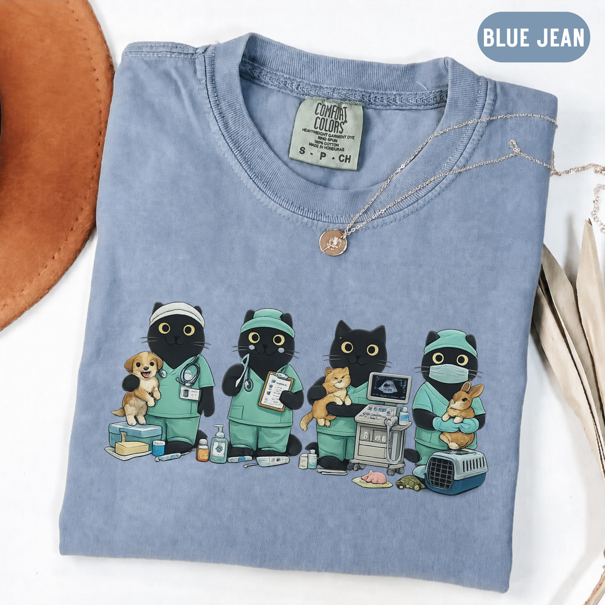 A folded light blue t-shirt with a graphic of four cartoon cats dressed as veterinarians, styled on a white surface with a brown hat and a gold necklace.