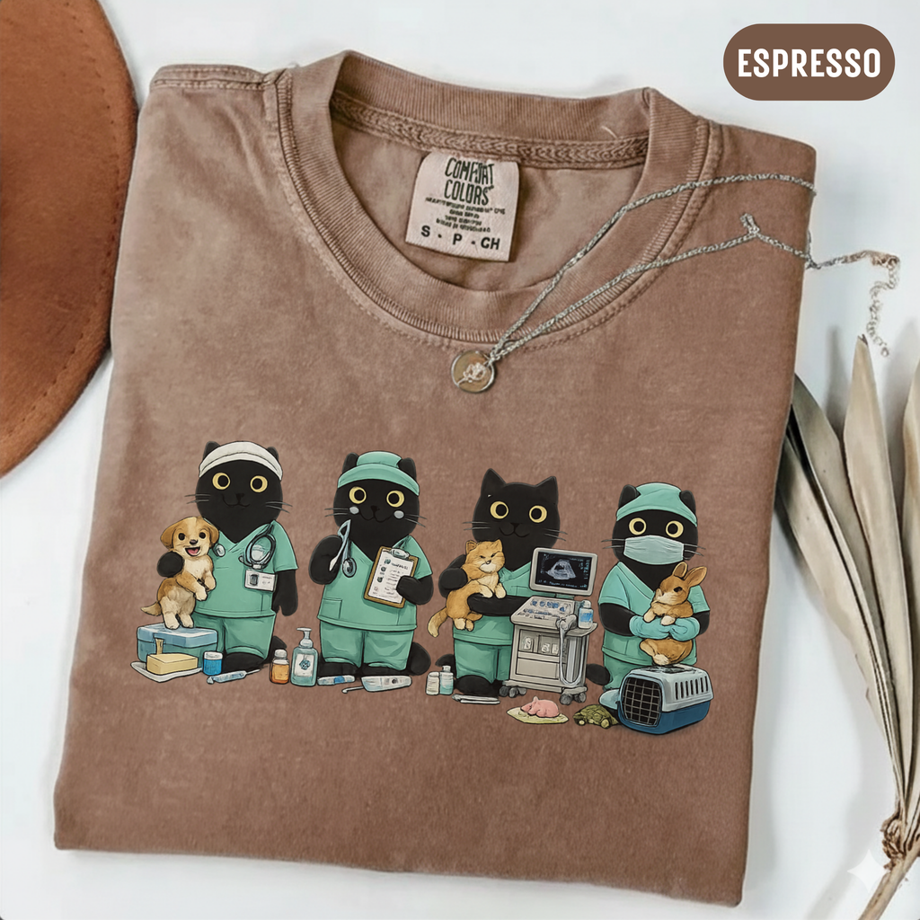A folded brown t-shirt with a graphic of four black cats dressed in green medical scrubs, each holding a small pet (dog, cat, rabbit, and bird). The shirt is styled on a white surface with a brown hat edge, a silver necklace, and dried leaves, with an 'ESPRESSO' label in the top right corner.