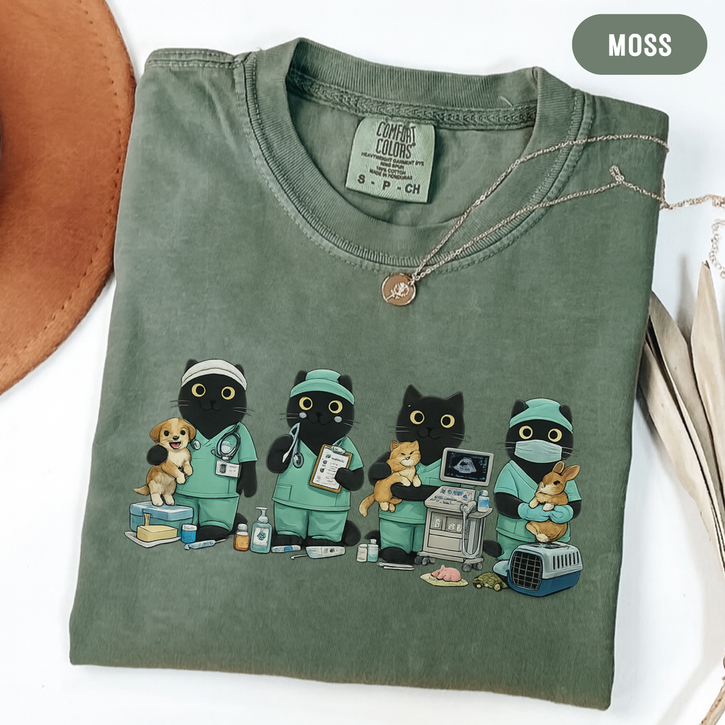 An olive green t-shirt with a graphic of four black cats dressed as veterinarians, styled flat on a white surface with a brown hat, a gold necklace, and a beige bag nearby. A 'MOSS' color label is visible in the top right corner.