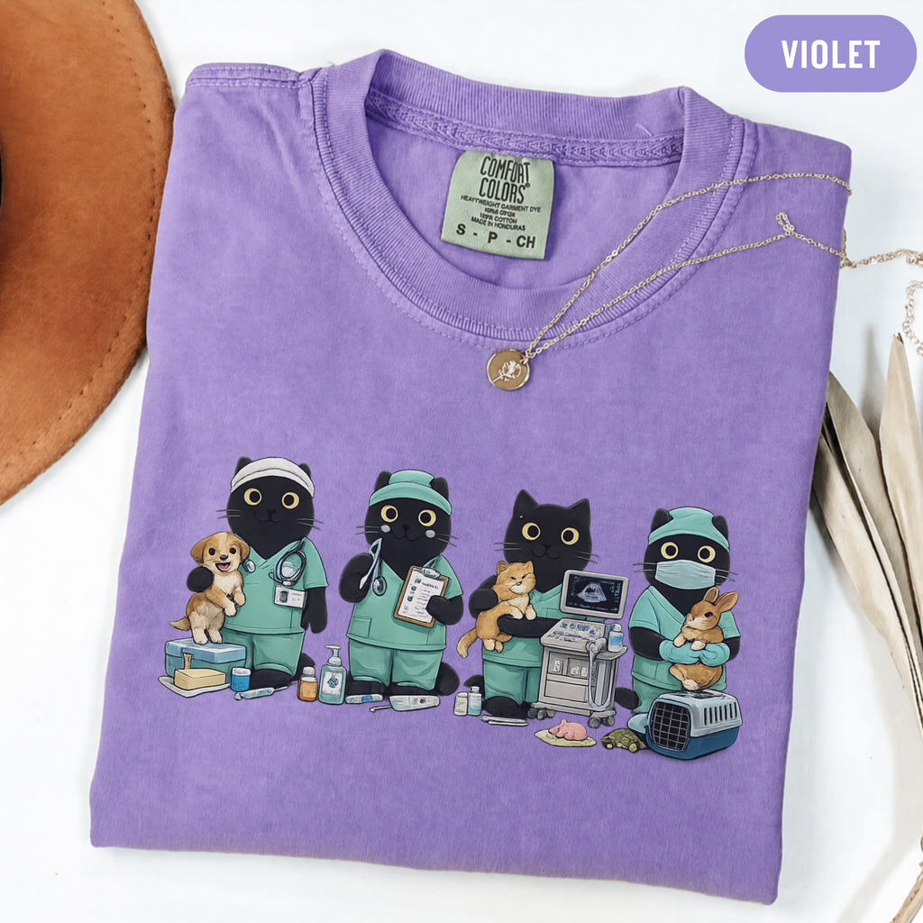 A folded purple t-shirt with a graphic of four black cats dressed in green medical scrubs, shown on a white background with a gold necklace draped over the collar and a purple 'VIOLET' label overlay.